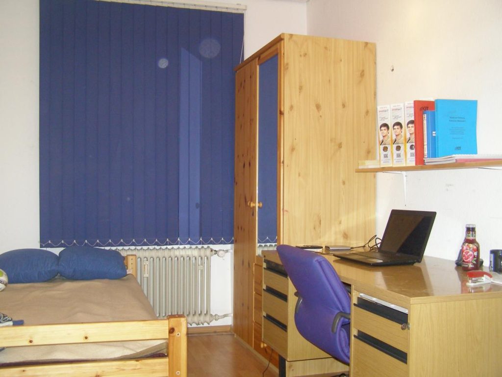 Kleines Zimmer mit Bett, Schrank und Schreibtisch.